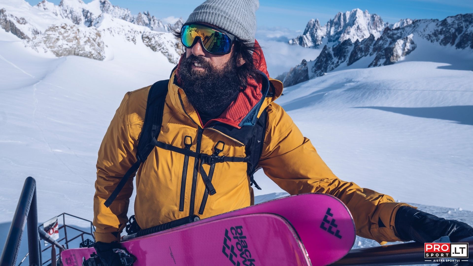 Barzdotas slidininkas, pasipuošęs stilingos kolekcijos Bogner slidinėjimo apranga, vienoje rankoje laikydamas slides ruošiasi nusileidimui